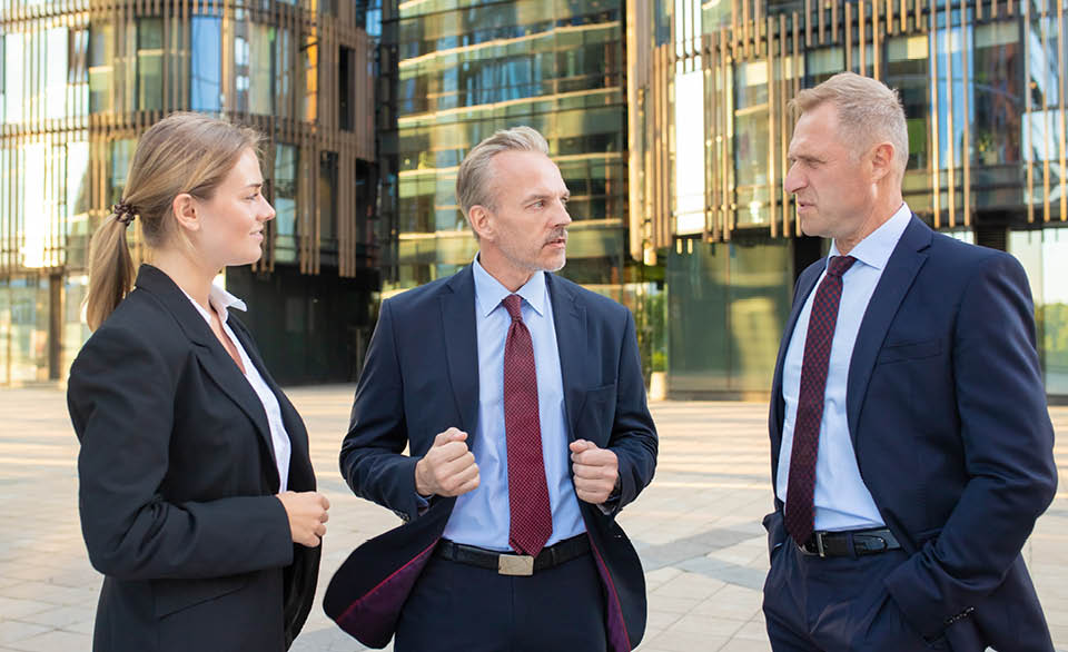 Three business professionals in suits talking outside a modern glass office building.