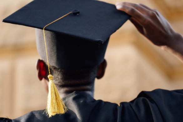 Close-up view from behind of a graduate wearing a black cap and gown adjusting a mortarboard with a gold tassel, standing outdoors near a brick building.