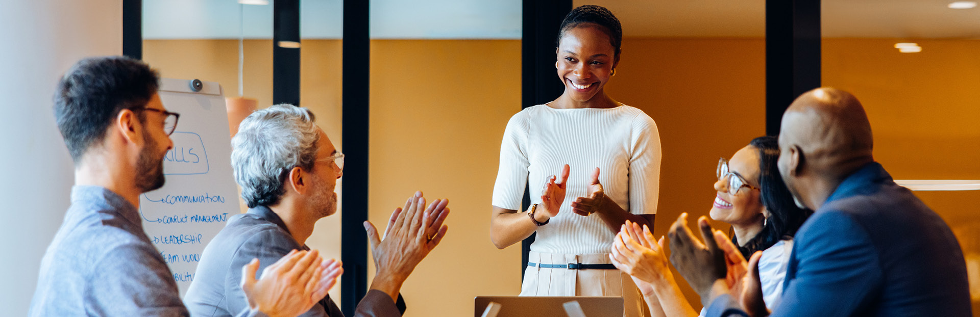 A group of professionals gathered around a conference table applaud while one person stands at the front of the room during a meeting or presentation.