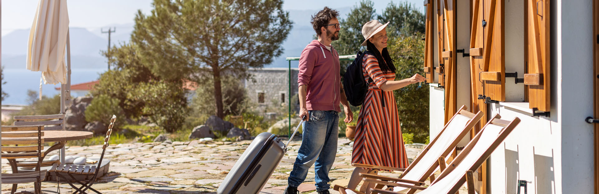 Travelers with luggage arrive at a vacation rental home with outdoor seating and deck chairs.