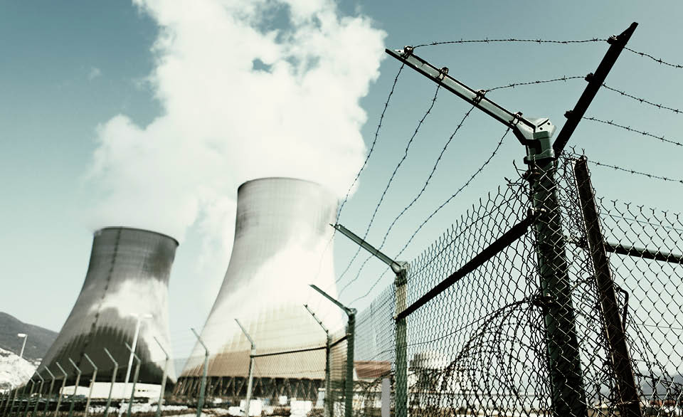 Cooling towers at a nuclear power plant behind a barbed wire fence under a cloudy sky.