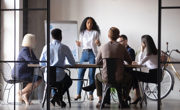Person standing and presenting to a small group seated around a conference table in a modern meeting room with a whiteboard nearby.
