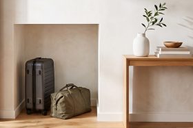 Black rolling suitcase and olive-green duffel bag placed in a small storage space next to a wooden table holding stacked books, a white vase with leafy branches, and a wooden bowl in a minimalist interior setting.