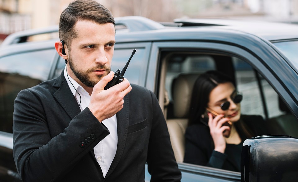 Person in an earpiece speaks into a two-way radio beside a parked SUV while another person uses a phone inside the vehicle.