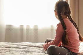 young girl sitting on her bed looking out the window.