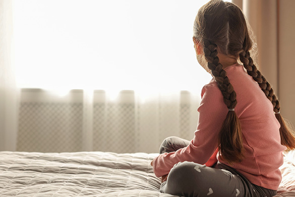 young girl sitting on her bed looking out the window.