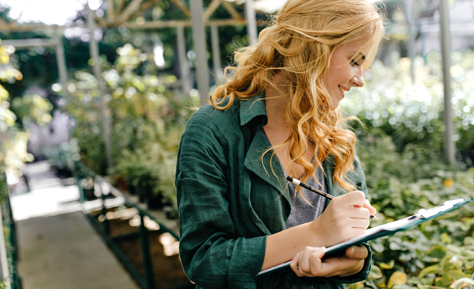 Person in a greenhouse records notes on a clipboard while inspecting plants.
