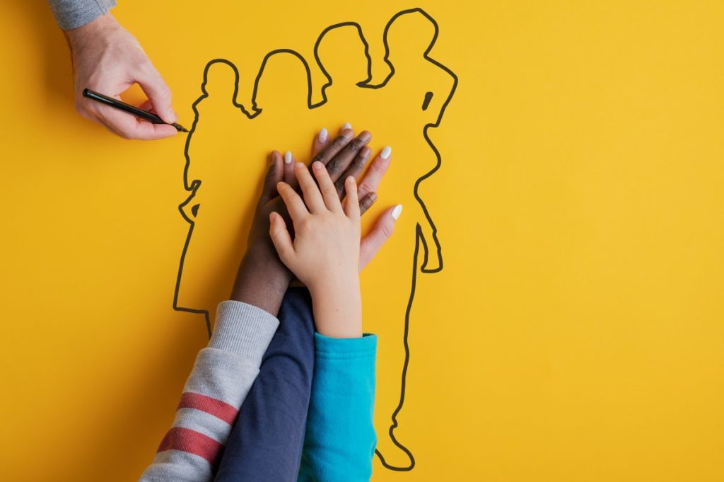 Small children's hands stacked together on a bright yellow background while a person draws the outline of a group of people around them, symbolizing teamwork, inclusion, diversity, and community collaboration.