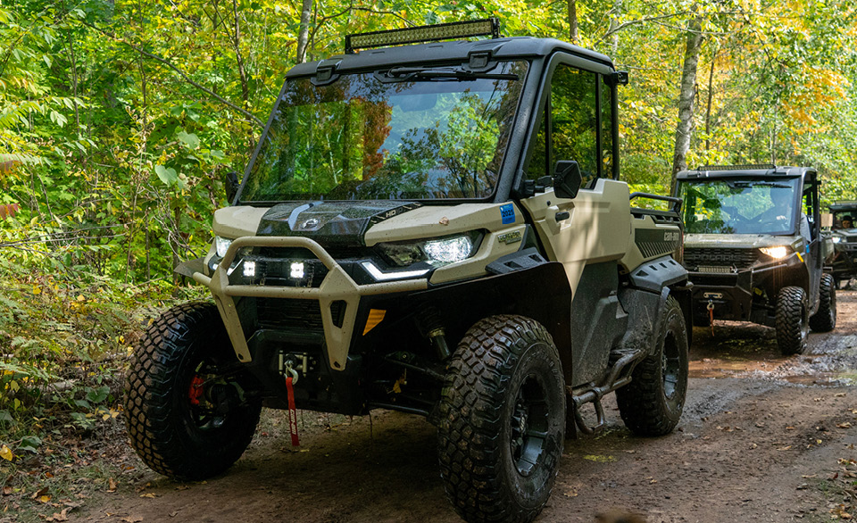 Two utility task vehicles drive along a wooded dirt trail.