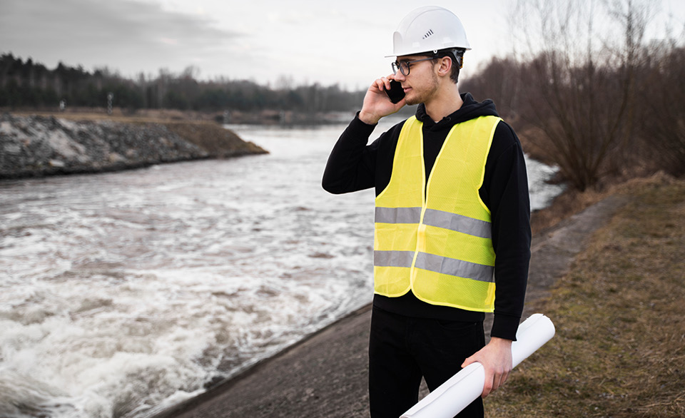 Person in a hard hat and safety vest uses a phone near a flowing waterway while holding rolled plans.